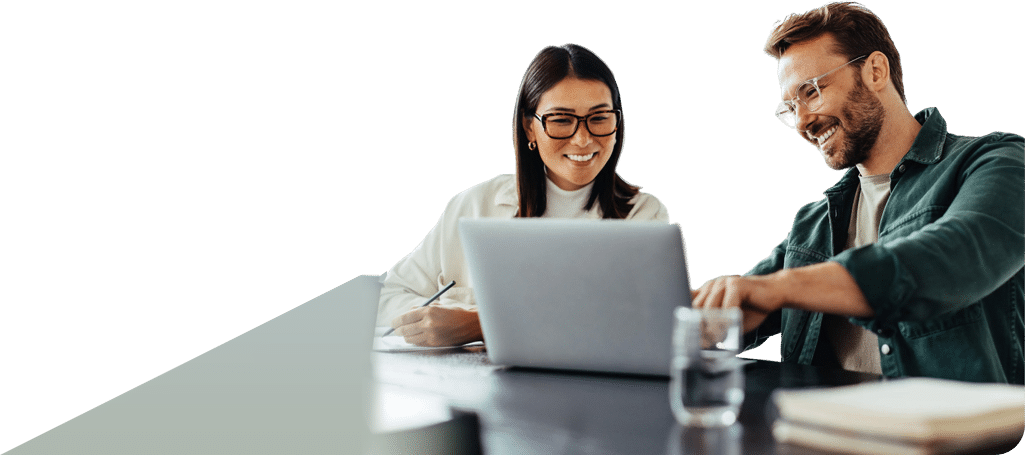 A man and woman with glasses smile while looking at a laptop together, with the man pointing at the screen and the woman holding a pen over some papers.