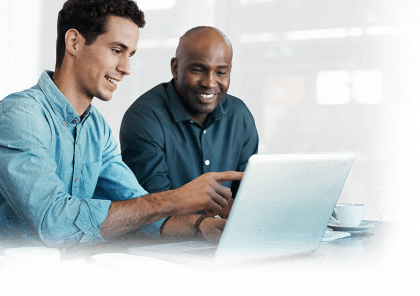 Two men are smiling and looking at a laptop screen together, with one man pointing at the display.