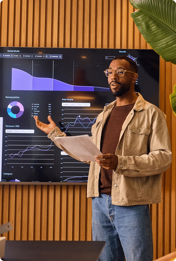 A man with glasses and a tan jacket stands in front of a large screen displaying data charts and analytics, gesturing toward the information while giving a presentation.