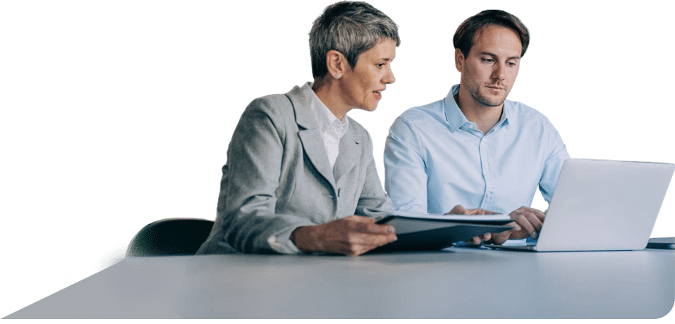 A woman with short grey hair in a blazer and a man in a light blue shirt sit side-by-side at a desk, focused on a laptop screen while reviewing documents.