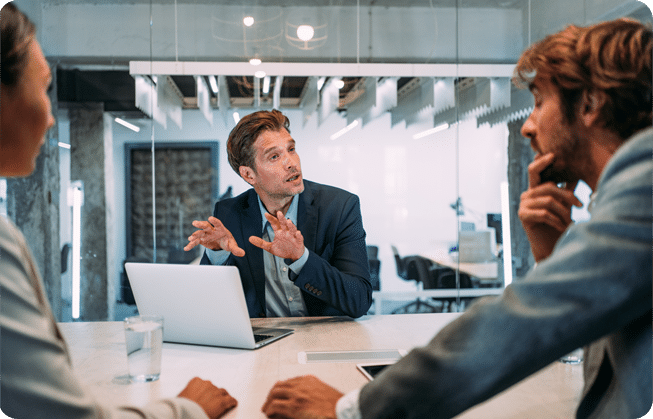 Three people in a modern office meeting, with one speaking and gesturing while others listen around a conference table.