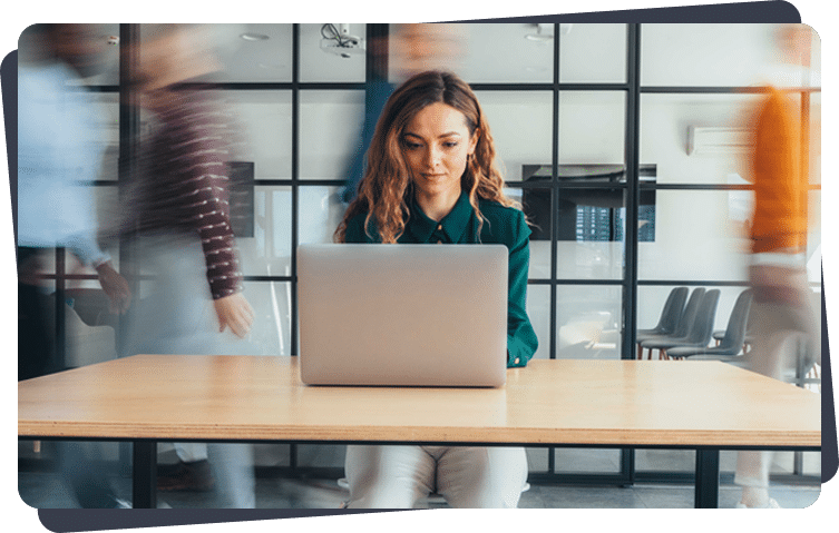 Person working on a laptop at a wooden table in a busy office with people walking past in the background.