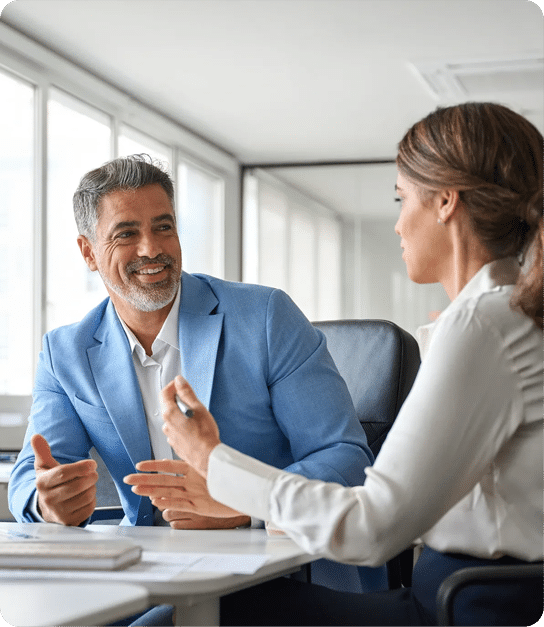 A man in a blue blazer and a woman in a white blouse sit across from each other at a desk, engaging in a friendly and professional conversation.