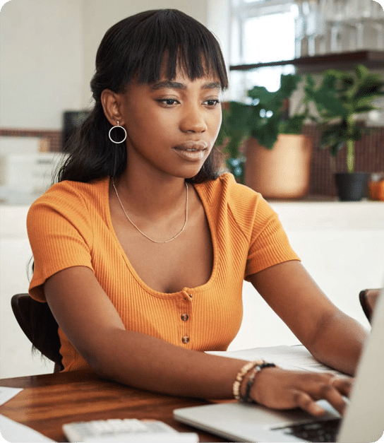 A person in an orange short‑sleeved shirt, wearing hoop earrings and bracelets, working on a laptop at a table in a cozy home workspace with plants and shelves in the background.