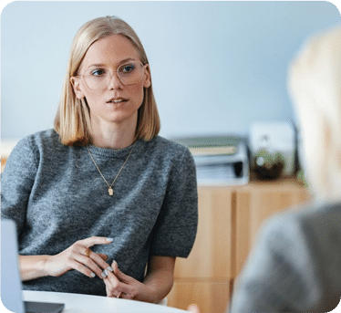 A woman with blonde hair and glasses wearing a grey sweater sits at a desk, looking attentively at someone off-camera during a conversation.