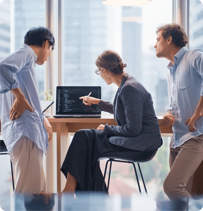 Three colleagues—two standing and one seated—collaborate in a modern office, focusing on a laptop screen as the woman points to lines of code on the display.